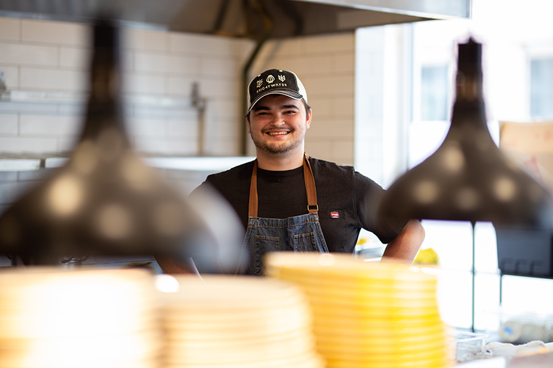 Smiling sous chef wearing black shirt, denim apron and black baseball cap that says Brightwater
