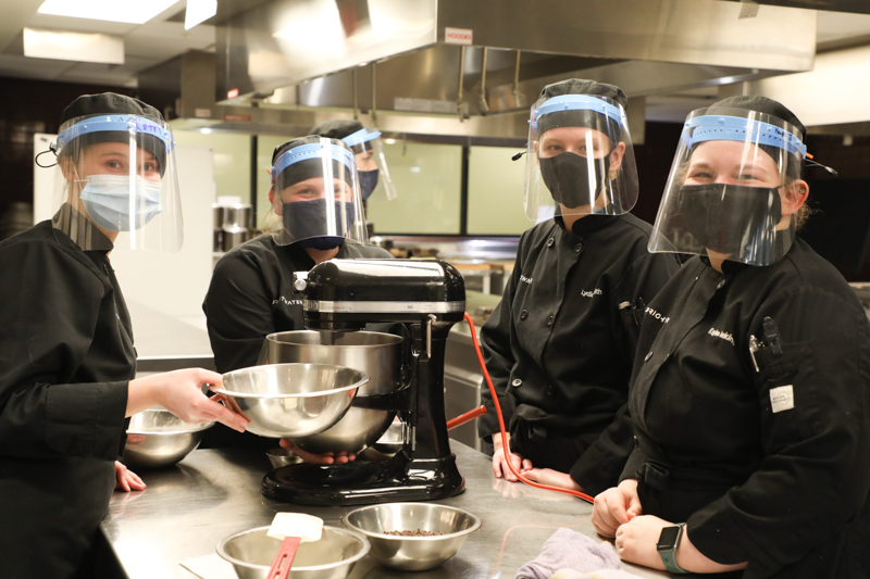 Four culinary students wearing black coats and face masks looking at camera