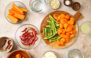 Overhead view of assorted peppers, carrots, garlic, onions, and seasonings arranged on a countertop for homemade hot sauce preparation.