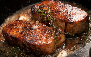 Close-up of two seared pork chops cooking in a skillet with garlic, herbs, and sizzling butter.