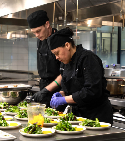 Two culinary students in black chef uniforms and hats carefully plate salads in a professional teaching kitchen. One student arranges ingredients on multiple white plates while another assists nearby. Stainless steel countertops, commercial cooking equipment, and prepared dishes create an active, hands-on food preparation environment.
