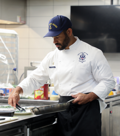 A chef in a white chef coat and blue cap prepares food at a stovetop in a commercial teaching kitchen. Wearing a U.S. Coast Guard uniform chef jacket, he focuses on cooking in a skillet while surrounded by professional kitchen equipment and ingredients, highlighting hands-on culinary training.